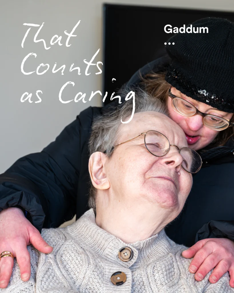 A woman with down syndrome standing behind a woman, presumably a loved one. She lovingly places her hands on her shoulders.