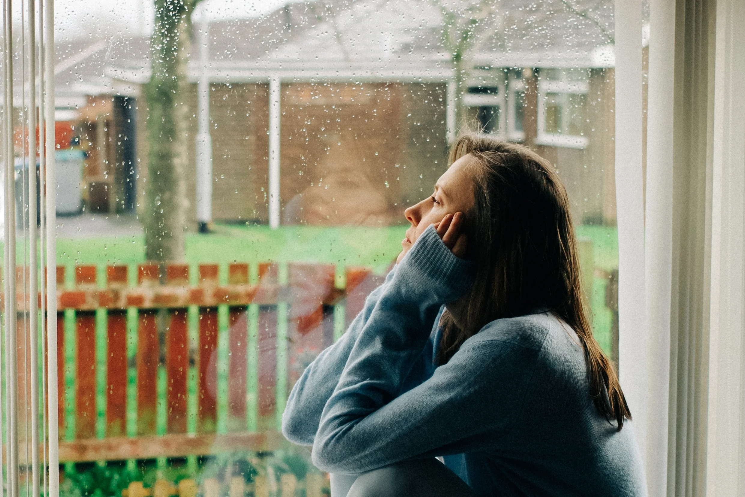 Thoughtful,Sad,Woman,Sitting,On,A,Windowsill,With,A,Reflection